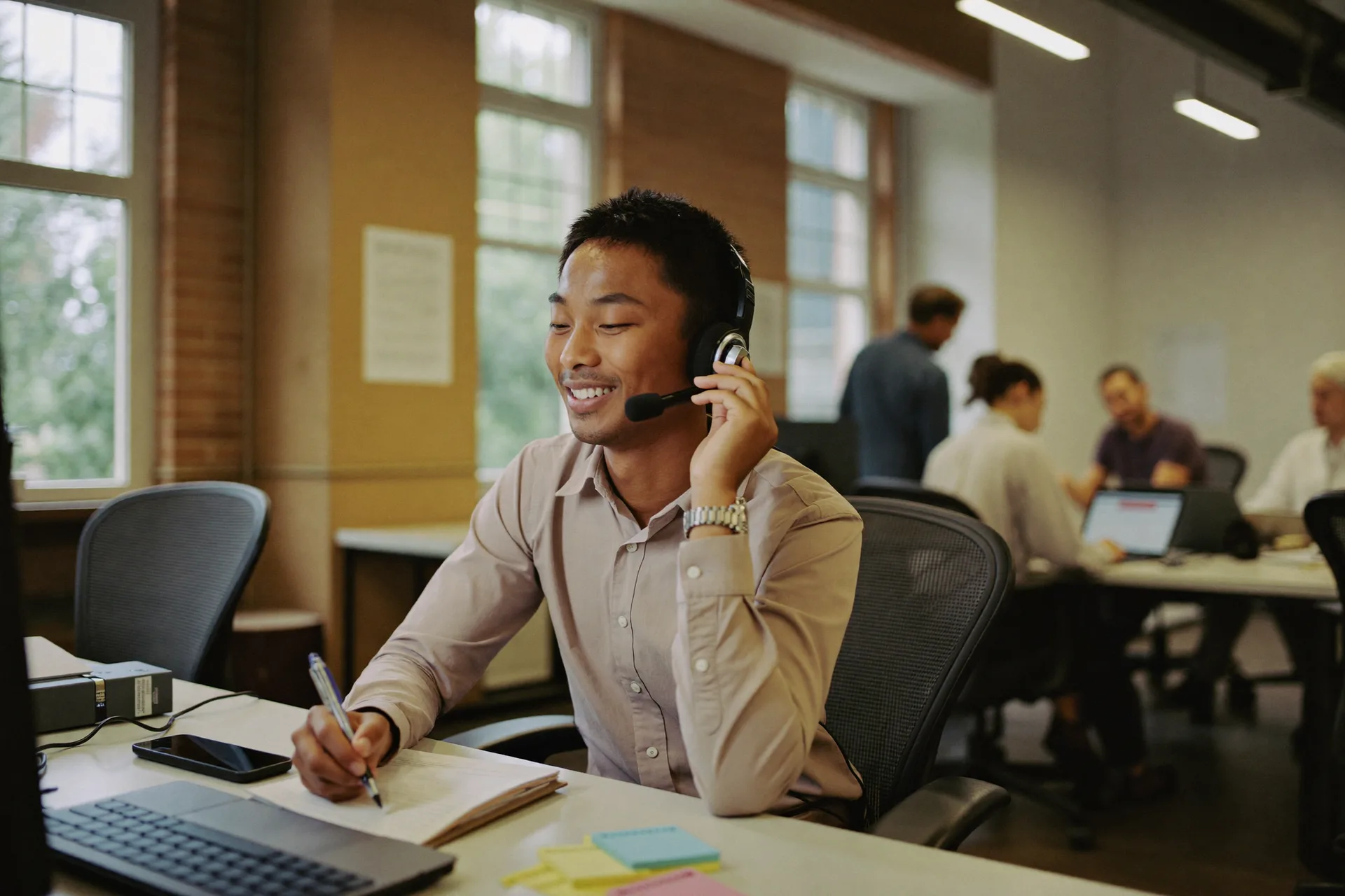 A man on a headset at his desk in a shared office, taking notes as he talks